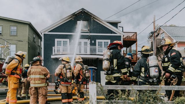Image of the playhouse on fire. Firefighters are spraying water into the top floor window. The upper floor is charred and smoke is in the air.
