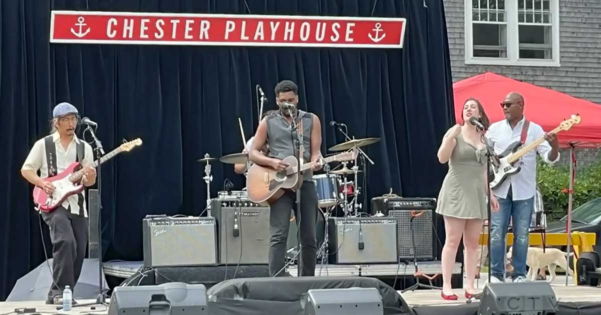 Image of a band on a stage outdoors. One Asian male, two African Nova Scotian male, and one Caucasian male. Black curtain behind them with red and white sign that reads Chester Playhouse.