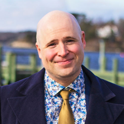 Image of Caucasian male. He is outside, facing the camera and smiling. He is bald, clean shaven and wearing a dark blue jacket, with blue and yellow floral shirt and yellow tie,