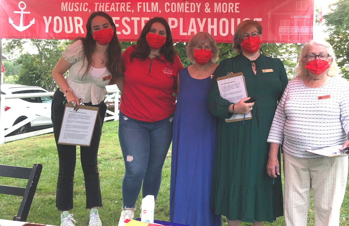 A colour photo of five women standing outside under a red tarp. They are all wearing red facemasks. wo are holding clipboards. A banner behind them reads Music, Theatre, Film, Comedy and More Your Chester Playhouse