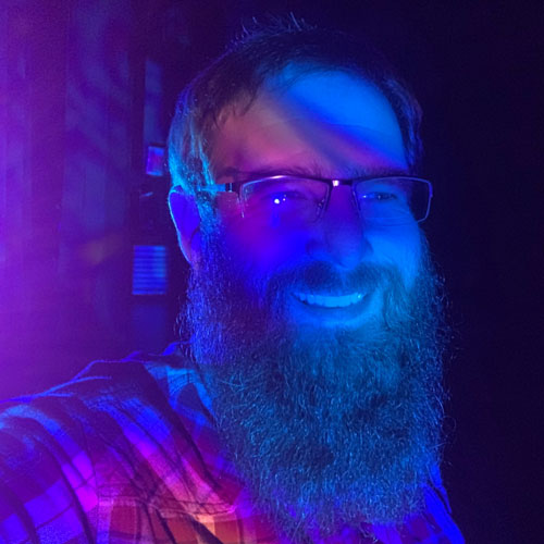 Image of young Caucasian man, with short dark hair and beard, He is wearing glasses and a plaid shirt. He is looking at the camera and smiling. He is bathed in purple light.