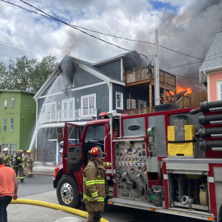 Image of the playhouse fire. Firefighters are shooting water into the third-floor window. Smoke is billowing out and flames can be seen at the rear of the building.