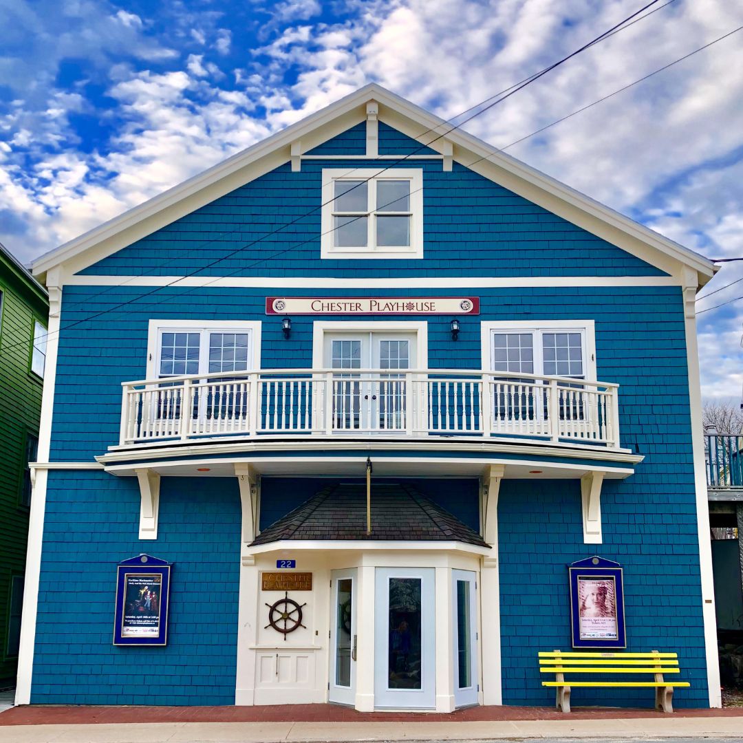 A colour image of the playhouse. A Three story building, blue siding, white balcony on the second floor. A yellow bench sits outside.